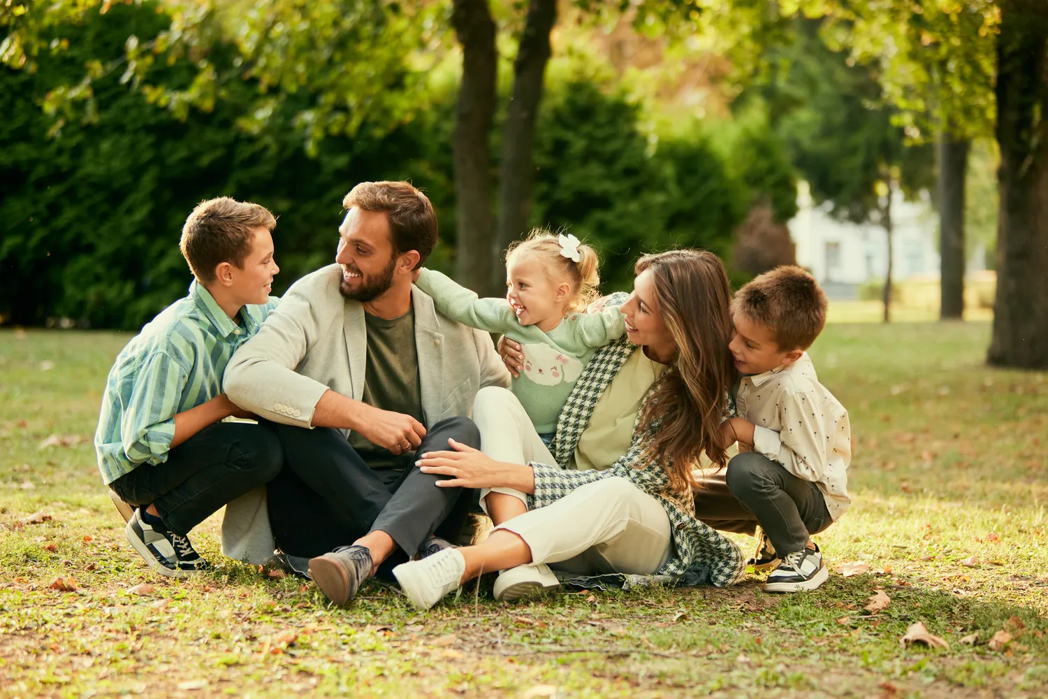 Herzensnah by Tanita Englisch -  Familiäre Unterstützung - Eine große Familie, bestehend aus zwei Erwachsenen und vier Kindern, sitzt zusammen auf der Wiese in einem Park, lächelt und unterhält sich miteinander.