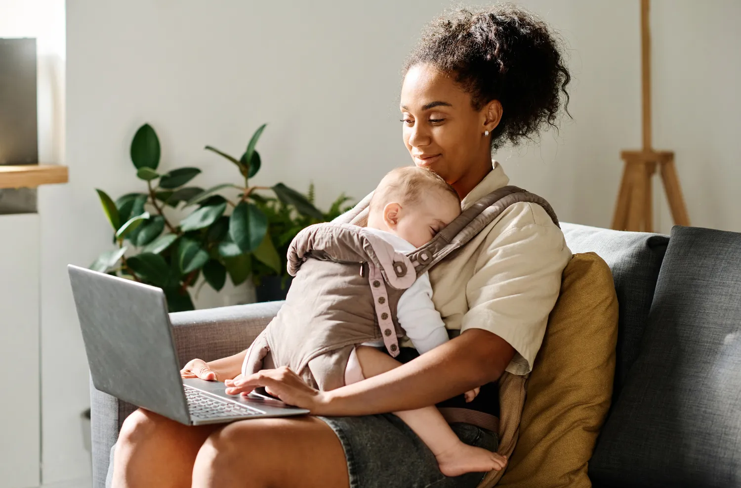 Herzensnah by Tanita Englisch -  Familiäre Unterstützung - Eine junge Frau sitzt mit einem Baby in einer Babytrage auf einem Sofa und tippt zu Hause auf ihrem Laptop.