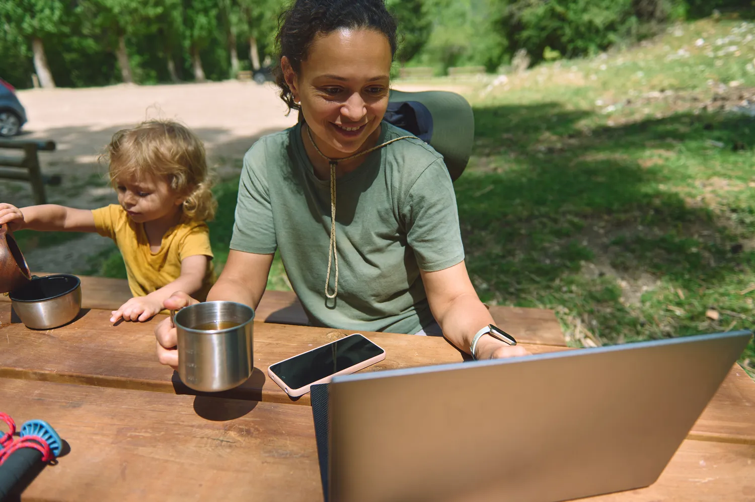 Herzensnah by Tanita Englisch -  Familiäre Unterstützung - Eine Frau und ein kleines Kind sitzen im Freien an einem Holztisch und schauen beide auf einen Laptop-Bildschirm. Ein Smartphone und ein Metallbecher stehen ebenfalls auf dem Tisch.